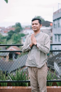 Genç bir Asyalı adam saygıdeğer bir selamlama sergiliyor ve namaste bir jest yapıyor. Arkasında binalar ve yeşilliklerle bir şehir ortamında kültür, saygı ve huzur duygusu taşıyor..