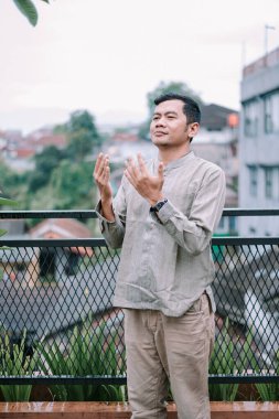 Balkonda el kaldırarak dua eden genç bir müslüman. Şehir manzarasında manevi bir minnettarlık ve bağlılık yaşıyor. Dini uygulamalarda inanç, inanç ve farkındalık göstergesi.