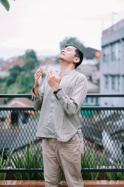 Geleneksel giysiler giymiş bir adam, bir balkonda dua ediyor, gözleri huzurlu bir şekilde dalgın dalgın bakıyor. Şehir mimarisi ve bulutlu gökyüzü bu bağlılık ve ruhsal bağlantı anı için sakin bir zemin oluşturur..