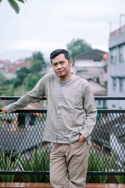 Düşünceli bir Asyalı adam terasta duruyor, korkuluklara yaslanıyor, doğrudan kameraya bakıyor. Normal bir şekilde bej pantolon ve desenli bir gömlek giyiyor, bir eli de cebinde. Şehir onun arkasından yayılıyor, yaratıyor.