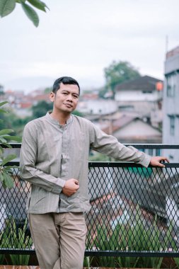 Düşünceli bir adam bir balkonda dikilip, kendinden emin bir şekilde ufka bakarken resmedilir. Gündelik giysiler giymiş, şehir manzarasının arka planında yer alıyor. Bu, kentsel bir ortamda rahatlatıcı bir düşünce anı yakalar..