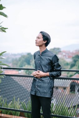 Genç bir adam çatıdaki bir balkonda, şehir manzarasına bakarak dalgın dalgın duruyor. Huzurlu ifade, bir anlık tefekkür ve huzuru yansıtır. Manzaranın ve temiz havanın tadını çıkarır..