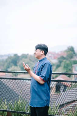 Geleneksel giyinmiş sakin bir Asyalı balkonda dua ederken ellerini kaldırmış duruyor. Dindar bir ibadetin, maneviyatın ve huzurlu bir düşüncenin görüntüsünü yakalıyor. Ayar yansıma için sakin bir arkaplan sağlar.