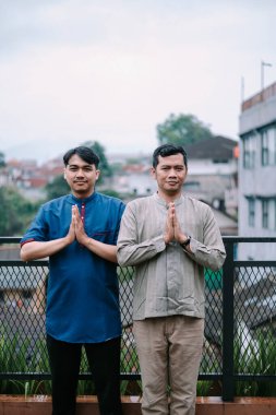 Geleneksel giysili iki dost canlısı Asyalı ellerinde saygılı bir selamlama jesti ile dışarıda poz veriyorlar. Şehir manzarası bulanık bir arkaplan sağlar ve resme bağlam ekler. İnanç, saygı ve kültürel gelenekleri temsil eder..