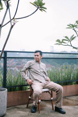 Güneydoğu Asyalı bir adam dışarıda bir sandalyede oturuyor, muhtemelen bir çatı ya da balkonda, manzaranın tadını çıkarıyor ve düşünüyor. Arka planda, sakin ve huzurlu bir atmosfer yaratan bir şehir silüeti ve yemyeşil yapraklar yer alıyor. Adam rahat görünüyor.