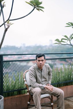 Düşünceli bir adam çatıdaki sandalyede oturur ve ötesindeki manzarayı seyreder. Sakin dış mekan ve rahat duruş sakin bir tefekkür hissi yaratır. Resim modern mimari ve doğal elementlerin bir karışımını vurguluyor.