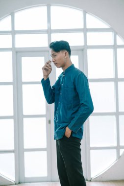 Şık mavi gömlekli düşünceli bir genç adam parlak bir pencerenin yanında düşünceli bir şekilde içeride poz veriyor. Onun eli yüzüne doğru hareket eder, düşünceli atmosferi artırır. Sahne, iç gözlem anını yakalar..