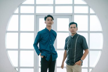 İki adam büyük bir pencerenin önünde sıradan gömlekler giyerek dikiliyorlar. Güven yayarlar ve modern ve dost canlısı görünürler. Bu görüntü moda, yaşam tarzı ve arkadaşlık temaları için uygundur..