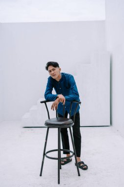 Parlak, bembeyaz bir odadaki tabureye yaslanan Asyalı genç bir adamın portresi. Minimalist ortam onun ifadesini ve kıyafetlerini vurguluyor, modern ve şık bir imaj yaratıyor..