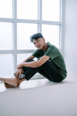 Genç adam büyük bir pencerenin yanında oturuyor, akıllı telefonunu tutuyor. Rahat ve huzurlu görünüyor, sıradan giyinmiş, yalın ayak. Bu iç mekan minimalist bir hava veriyor, yaşam tarzı kavramları için mükemmel..