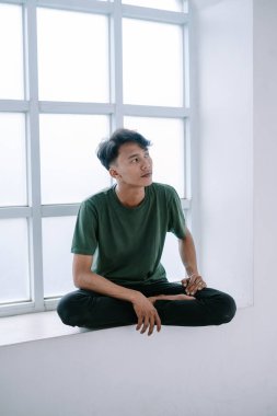 Asyalı bir adam, pencerenin pervazında bağdaş kurarak oturuyor. Yüzünde düşünceli ve barışçıl bir ifade var. Pencereden dışarı bakıyor. Bir anlık sakinlik ve yansıma izlenimi veriyor..