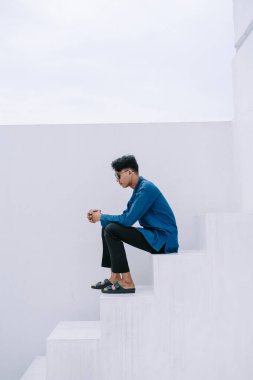 Düşünceli genç bir adam, derin düşüncelere dalmış, beyaz basamaklarda oturur. Modern bir bina cephesinin minimalist yapısı yalnızlık ve sessiz içgözlem duygusunu güçlendirir. Sıradan giyinmiş, güneş ışığıyla yıkanmış, gençlik ve gençlik katıyor.