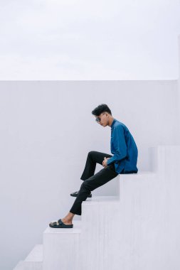 Gösterişli mavi gömlek ve koyu renk pantolon giymiş şık bir genç adam düşünceli bir şekilde beyaz basamaklara oturur. Görüntü, modern moda ve minimalist estetiğin bir karışımıdır. Güneş gözlüğü ve sandalet giyiyor ve düşünceli bir ruh hali içinde..