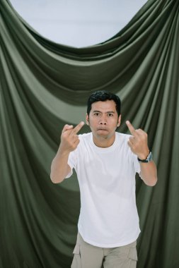 Beyaz tişörtlü bir adam iki eliyle tahrik edici bir jest yapar, örtülü bir arka plana karşı sert bir ifade sergiler. Görüntü güçlü öfke, meydan okuma ve hayal kırıklıklarını gösteriyor..