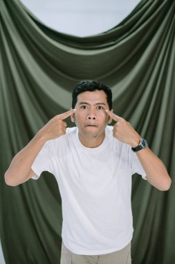 Asyalı bir adam, stüdyo ortamında nötr yeşil bir zemin üzerine kurulmuş, kafası karışık bir ifadeyle gözlerini işaret eder. Beyaz bir tişört ve haki rengi pantolon giyiyor, rahat ve uyumlu bir hava yaratıyor. Görüntü bir şaşkınlık anı yakalar.