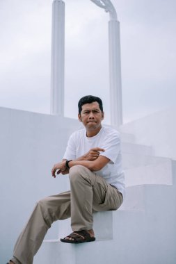 Bir adam, kentsel bir ortamda anıtsal sütunların yanındaki beyaz basamaklarda düşünceli bir şekilde oturur. Basit bir kıyafet giyiyor. Yüzündeki ifade derin düşüncelere daldığını ya da derin düşüncelere daldığını gösteriyor..