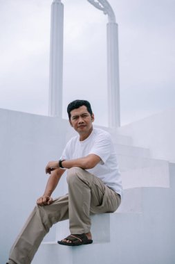 Beyaz merdivenlerde oturan dalgın bir adam, görünüşe göre bir anlık tefekkür içinde. Arka planda mimari unsurlarla gelişigüzel giyinmiş. Tarafsız tonlar ve bulutlu gökyüzü sakin ve huzurlu bir his veriyor..