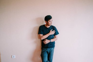 Sıradan giyinmiş düşünceli Asyalı bir adam düz bir duvara yaslanıyor. Başını çeviriyor, eli göğsünde dalgın ve dalgın görünüyor. Kopyalama alanına sahip minimalist biçim.