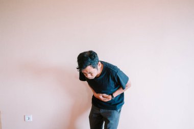 Bir adam karın ağrısı ya da hastalık belirtileri göstererek, karnını tutarak, rahatsız bir şekilde eğilir. Sağlık sorunları, sindirim sorunları veya genel fiziksel rahatsızlığı göstermek için idealdir..