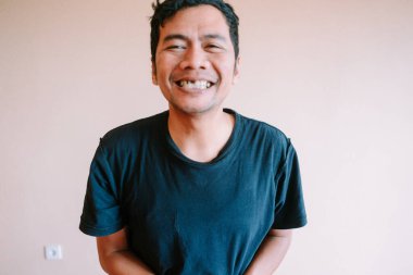 Neşeli Asyalı bir adam kapalı mekan portresi için gerçekten gülümsüyor. Gizli çekim, onun mutlu ve olumlu ifadesini düz bir duvara yansıtıyor. Günlük kıyafetleri ve rahat tavırları basit ve otantik bir yaşam tarzını yansıtıyor..