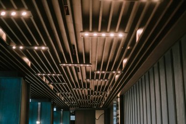 Paralel ahşap kirişleri ve aralıklı aydınlatma armatürleri olan modern bir mimari tavan. Tasarım, iç mekâna biraz zarafet katarak şık ve ritmik bir kalıp yaratıyor. Geçmişe uygun ya da modern tasarımı temsil eden elem