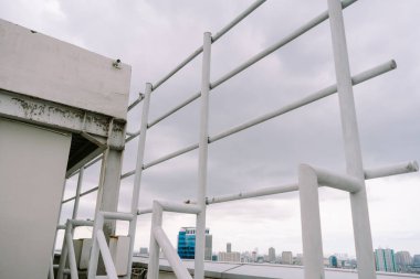Beyaz metal güvenlik korkuluklarından bakan panoramik şehir manzarası kasvetli bir gökyüzünün altında sergileniyor. Mimari ayrıntılar sanayi yapısı ile uzak şehir manzarası arasındaki zıtlığı vurguluyor. Özellikler bir gözlem bakış açısı, bir ileti 