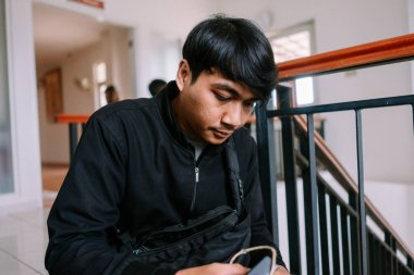 Genç bir adam kapalı bir merdivende otururken akıllı telefonunu kullanıyor. Askı çantalı siyah bir ceket giyiyor. Bu resim modern yaşam tarzını, iletişimi ve teknolojiyi betimliyor..