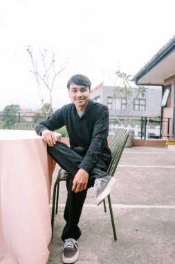 Siyah giysiler içinde gülümseyen genç bir adam, dışarıda bir sandalyede dinleniyor. Kendinden emin ve mutlu görünüyor. Sıradan ve dostane bir imaj çiziyor. Yumuşak açık hava ışığı portrenin misafirperver ve cana yakın doğasını geliştirir..