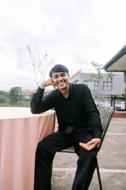 Parlak bir gülümsemesi olan neşeli genç bir adam dışarıda bir sandalyede oturuyor. Günlük giysiler giymiş ve mutluluk ve rahatlama hissi yayıyor. Ayarlar modern ve kentsel bir yaşam tarzını, kendine güvenen bir keyif anını temsil ediyor..