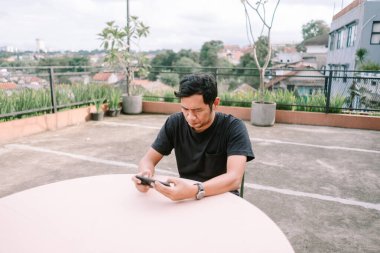 Çatıdaki terasta akıllı telefonunu kullanan Asyalı bir adam. Yaz havasının tadını çıkarıyor ve sosyal medya aracılığıyla başkalarıyla iletişim kuruyor. Arka planda şehrin ufuk çizgisi görünür..