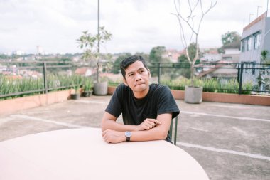 Düşünceli bir adam çatı katında bir masada oturuyor, düşüncelere dalmış. Şehir manzarası, bireyin sessiz tefekkürünü vurgulayan bulanık bir arka plan sağlar. Açık teras ayarları rahatlama ve yansımayı davet eder.