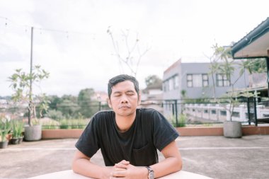 Huzurlu Asyalı bir adam kapalı gözlerle oturur, açık havada farkındalık ve meditasyon pratiği yapar. Şehir manzarası arasında iç huzur ve dengeli bir devlet arıyor. Çatı katı, düşünceli yansıma ve kişisel refah için sessiz bir kaçış sağlar.