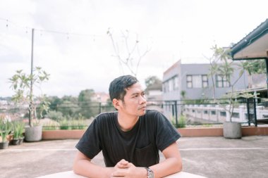 Asyalı bir adam terasta açık bir masada oturuyor, düşünceli bir şekilde uzaklığa bakıyor. Sahne, kentsel bir ortamda düşünceli ve yalnızlık hissi veriyor. Yansıma gibi çeşitli kavramları resmetmek için kullanılabilir.