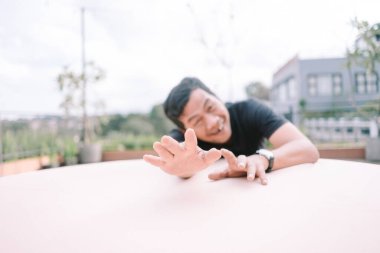 Neşeli Asyalı bir adam şakacı bir şekilde çatı katındaki terastan izleyiciye uzanıyor. Gülümseyen yüzü ve günlük kıyafetleri kaygısız ve cana yakın havasına katkıda bulunuyor. Açık hava manzarası, bulanık şehir manzarası ve yeşillikle birlikte, bir Sen 'i çağrıştırıyor.