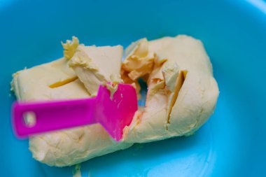 Tereyağının turkuaz bir kasede pembe bir spatulayla hazırlandığı yakın plan görüntüsü. Tatlıları hazırlamak için gerekli malzemelerin hazırlanmasını, canlı renklere sahip bir mutfak ortamını ve basit ev yapımı yemeklere odaklanmayı gösteriyor..
