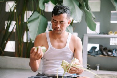 Bir adam bayram kutlaması için geleneksel pirinç köftesi ketupat hazırlıyor. Palmiye yapraklarını tipik elmas şeklinde dokuyor. Kültürel gelenek ve şenlikli yemek hazırlama ruhunu yansıtıyor..