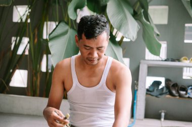 Kolsuz tişörtlü Asyalı bir adam açık havada bitki budamaya odaklanıyor. İşine odaklanmış, bahçesini sakin ve tropikal bir ortamda idare ediyor. Bir hobi, refah ve basit bir ev hayatını temsil eder..