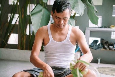 Asyalı bir adam geleneksel bir bayram kutlaması için palmiye yapraklarından ketupat dokuyor. Bu etkinlik, zengin kültür mirasını ve tatiller için süslemelerde kullanılan yerel becerileri gözler önüne seriyor. O özenle çalışıyor. 