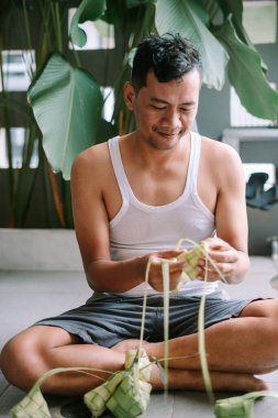 Bir adam, bayram kutlaması için geleneksel pirinç köftesi olan ketupları titizlikle dokuduğu görülüyor. Bereketli tropikal yaprakların arasında bağdaş kurmuş oturuyor. Kültürel ana doğal ve huzurlu bir dokunuş ekliyor. Karmaşık dokuma refleksi!