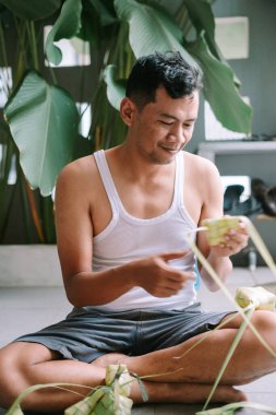 Bir adam bağdaş kurmuş, titizlikle palmiye yapraklarından ketupat dokuyor. Bu geleneksel etkinlik, kültür mirasını ve aşçılık sanatını temsil eden festival etkinliklerinin hayati bir parçasıdır. Odaklanmış ifadesi, gereken yetenek ve sabrı yansıtıyor.