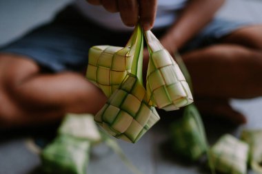 Elinde taze yapılmış ketupat tutan birini yakından çek. Örülmüş palmiye yaprağı kesesine doldurulmuş pirinçten yapılmış geleneksel bir yemek. Bu yemek birçok Güneydoğu Asya ülkesinde popülerdir ve bayram tatillerinde sıklıkla hizmet vermektedir..
