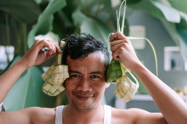 Gülümseyen Asyalı bir adam geleneksel pirinç keki tabağı Ketupat 'la oyun oynayarak Kurban Bayramı' nı kutluyor. Neşeli bir olayı sembolize eden örülmüş hindistan cevizi yaprakları ve pirinç birlikteliğin kültürel bir sembolünü oluşturuyor..