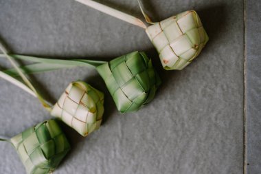 Titizlikle örülmüş ketupanın yukarıdan çekimi, geleneksel pirinç köftesi, dokulu bir arka planda. Mutfak mirasını temsil eden bu lezzetler, Kurban Bayramı gibi Güneydoğu Asya kutlamalarının kültürel zenginliğini somutlaştırıyor..