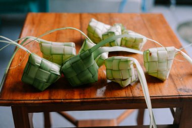 Geleneksel pirinç hamur tatlısı olan ketupat 'ın yakın çekimi, Güneydoğu Asya' daki bayram kutlamaları sırasında klasik ahşap bir masada yapılıyor. El yapımı bir lezzet, dokunmuş palmiye yaprakları buharda pişmiş pirinç içeren elmas bir şekil oluşturuyor..