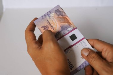 Yakın plan çekimde, bir kişinin elleri düzgünce istiflenmiş bir yığın Endonezya Rupia banknotunu tutuyor. Konferansta para biriminin dokusu ve ayrıntıları, finans, ekonomi ve parasal işlemler konuları ele alınıyor. Görüntü bir sen çağrıştırıyor.