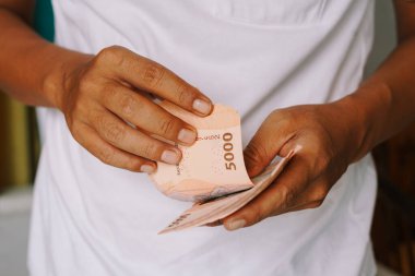 Beş bin Endonezya rupia banknotunu sayarak el ele tutuşun. Görüntü para işleme, finans, zenginlik ve ekonomiyi vurguluyor. Sahne, refah ve modern hayatta finansal planlamanın önemini gösteriyor..