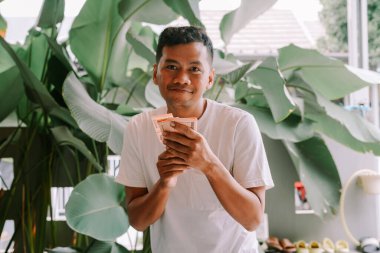 Gülümseyen ve elinde Endonezya Rupiası banknotları tutan Asyalı genç bir adamın portresi. O, yemyeşil yapraklarla çevrilidir. Zenginlik, yatırım, mali başarı ve refah kavramı.