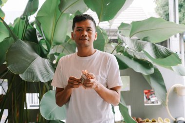 Çiçekli bir bahçede duran gülümseyen bir adam titizlikle kağıt paraları sayıyor. Tropikal bitkilerle çevrelenmiş halde, finansal refahı, yatırım başarısını ve girişimciliğin ödüllerini temsil ediyor. Zenginliğin ve bolluğun sembolü..