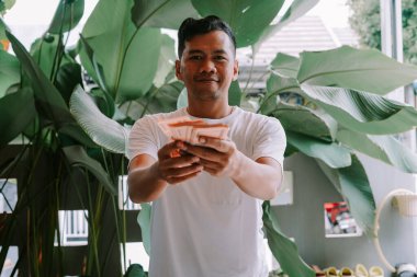 Sıradan beyaz tişörtlü, elinde banknot hayranı olan mutlu bir Asyalı. Gülümsüyor ve büyük, yemyeşil bitkilerle çevrili para birimini teklif ediyor. Zenginliği, finansal istikrarı veya başarılı bir yatırım fırsatını temsil eden büyük bir imaj.