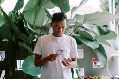 Asyalı bir adam Endonezya Rupiah banknotlarını titizlikle sayıyor. Açık havada, yemyeşil tropikal yapraklarla çevrili. Mali durumunu idare etmeye odaklanmış görünüyor, belki de kazancını hesaplamaya ya da değerlendirmeye..